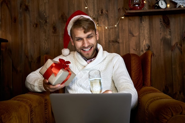 Gelukkige jonge man draagt een kerstmanhoed en zegt een toast. hij viert een virtueel nieuwjaarsfeest met een laptop. hij houdt een glas champagne en een kerstcadeau vast.