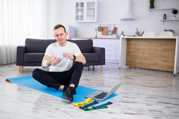 Gelukkige jonge man drinkt water na online training. Thuis sporten, gezonde levensstijl