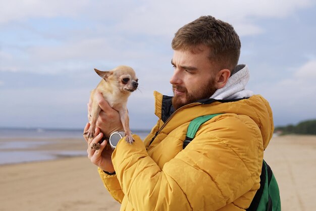 Foto gelukkige jonge man loopt met zijn kleine huisdier chihuahua of speelgoed terrier hond op het strand houden puppy op