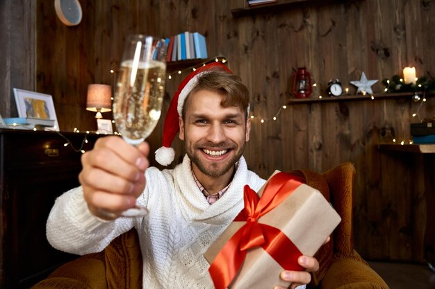 Gelukkige jonge man met een kerstmanhoed die een kerstcadeau vasthoudt en naar de camera kijkt opgewonden man die champagne drinkt met een virtuele chat met een vriend die het nieuwe jaar viert online feest op afstand webcam view