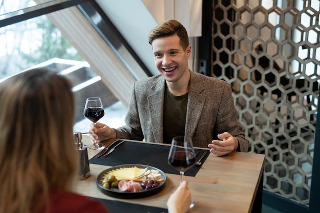 Gelukkige jonge man met glas rode wijn zittend aan tafel voor zijn vriendin en toast maken tijdens een romantisch diner in restaurant