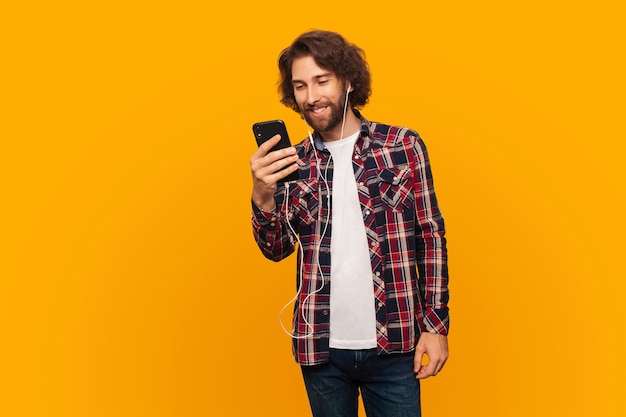 Gelukkige jonge man met krullend haar in shirt luistert naar muziek in koptelefoon en houdt smartphone vast