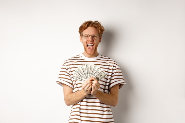 Gelukkige jonge man met rood haar die dollars toont, geld wint en schreeuwt van geluk, prijzengeld vasthoudt, staande op een witte achtergrond