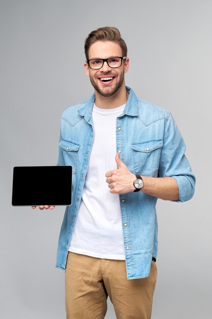 Gelukkige jonge mens die jeansoverhemd draagt dat en het stootkussen van tabletpc over studio grijze muur houdt houdt