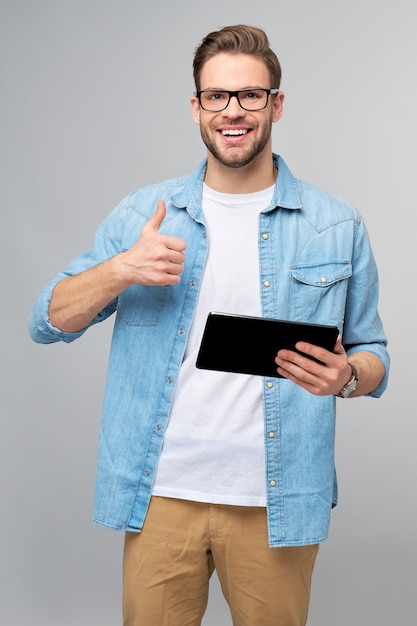 Gelukkige jonge mens die jeansoverhemd draagt dat en tablet over studio grijze muur draagt