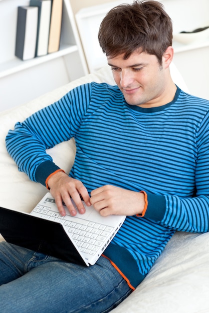 Gelukkige jonge mens die zijn laptop met behulp van die op de bank liggen