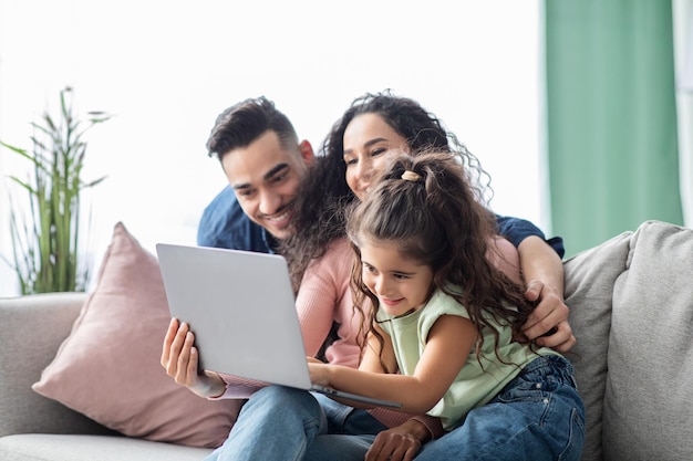 Gelukkige jonge midden-oosterse familie van drie tijd doorbrengen met laptop thuis, vrolijke arabische ouders en hun dochtertje met behulp van computer voor online winkelen of surfen op internet, close-up