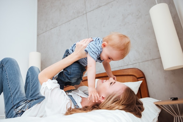 Gelukkige jonge moeder die thuis speelt en plezier heeft met haar zoontje op bed