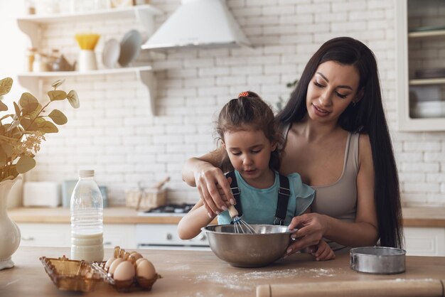 Gelukkige jonge moeder en klein babymeisje bereiden deeg en hebben plezier terwijl ze thuis koekjes maken in de keuken