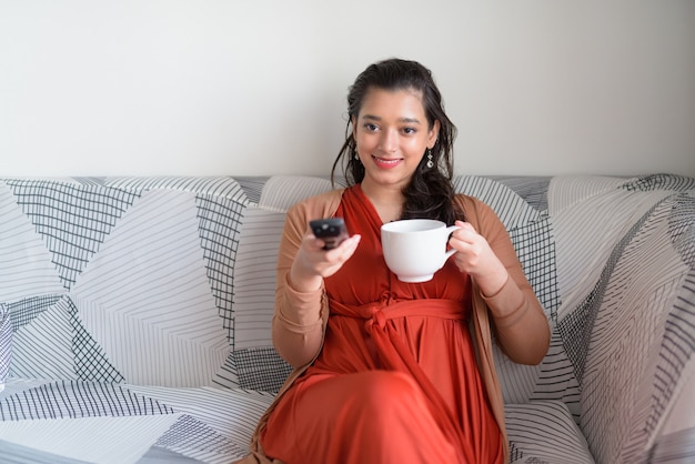 Gelukkige jonge mooie Indische vrouw die koffie drinken terwijl thuis het letten van op TV