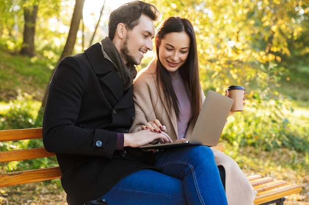 gelukkige jonge mooie verliefde paar poseren buiten wandelen in de natuur van het park zittend op de bank met behulp van laptopcomputer.