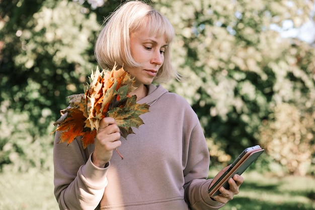 Gelukkige jonge mooie vrouw die buiten in het herfstpark loopt met behulp van chatten op de mobiele telefoon