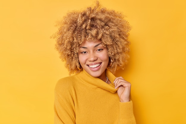 Gelukkige jonge mooie vrouw lacht tandjes rekt kraag van trui voelt goed drukt positieve emoties geïsoleerd over levendige gele achtergrond. Vrolijk vrouwelijk model met krullend borstelig haar.