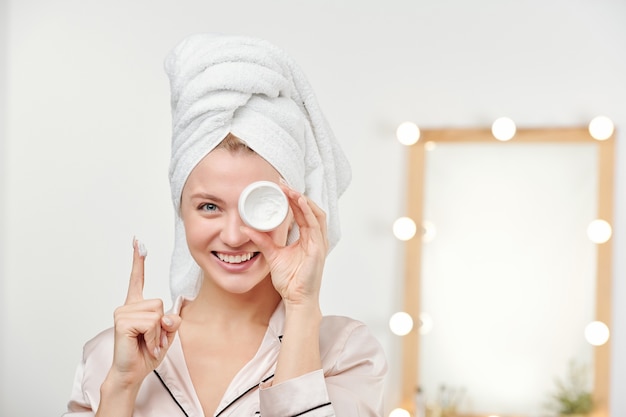 Gelukkige jonge mooie vrouw met een witte handdoek op het hoofd die haar gezichtscrème toont terwijl ze het door het linkeroog vasthoudt