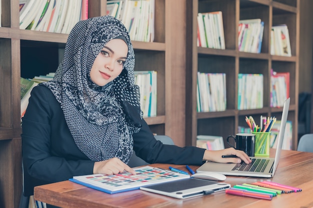 Gelukkige jonge moslim creatieve ontwerpervrouw die de steekproeven en laptop van het kleurenpalet voor boekenrek gebruiken.