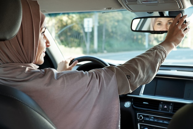 Gelukkige jonge moslimzakenvrouw in hijab die in de spiegel kijkt terwijl ze aan het stuur zit en auto rijdt