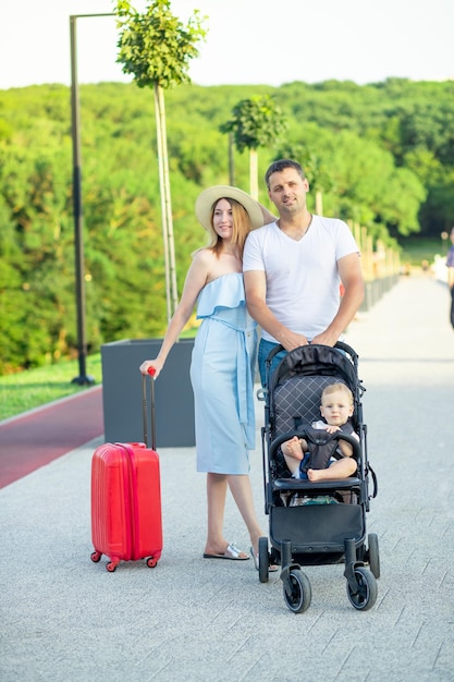 Gelukkige jonge ouders, mama en papa met een rode koffer en een baby in een kinderwagen, gaan in de zomer glimlachend op vakantie