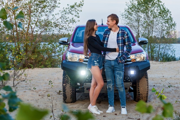 Gelukkige jonge paar verliefd glimlachend en knuffelen naast rode auto op het strand in de buurt van het meer. Jong koppel knuffels in de buurt van de auto.