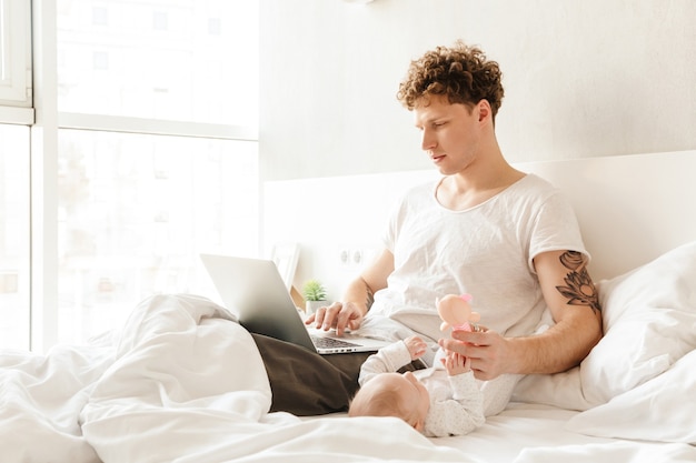 Gelukkige jonge vader die met zijn zoontje speelt terwijl hij op een laptopcomputer werkt en in bed ligt