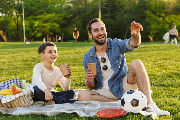 Gelukkige jonge vader met een picknick met zijn zoontje in het park, drinkend uit een beker