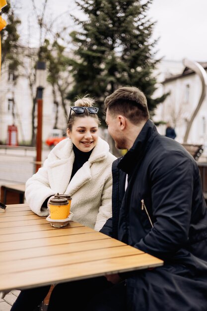 Gelukkige jonge verliefde paar in bovenkleding zittend aan een tafel buiten en koffie drinken Ontspannen in de open lucht