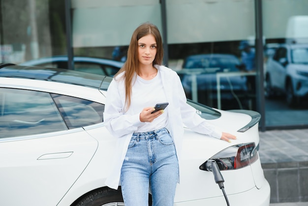 Gelukkige jonge volwassen vrouw die lacht en wegkijkt terwijl ze de auto-accu oplaadt van een klein openbaar station dat in de buurt van een elektrische auto staat