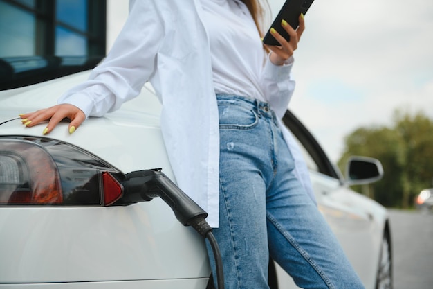 Gelukkige jonge volwassen vrouw die lacht en wegkijkt terwijl ze de auto-accu oplaadt van een klein openbaar station dat in de buurt van een elektrische auto staat