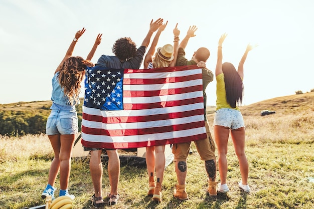 Gelukkige jonge vrienden genieten van een zonnige dag in de natuur. Ze kijken naar de zon die de Amerikaanse vlag vasthoudt en groet, blij om samen te zijn.