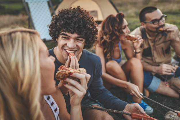 Gelukkige jonge vrienden genieten van een zonnige dag op de berg. Ze lachen en eten gegrilde worstjes bij de tent.