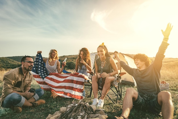 Gelukkige jonge vrienden Vierde juli-viering en geniet van een zonnige dag op de berg. Ze houden de Amerikaanse vlag vast, lachen en roosteren met bierflesjes in de buurt van de tent.