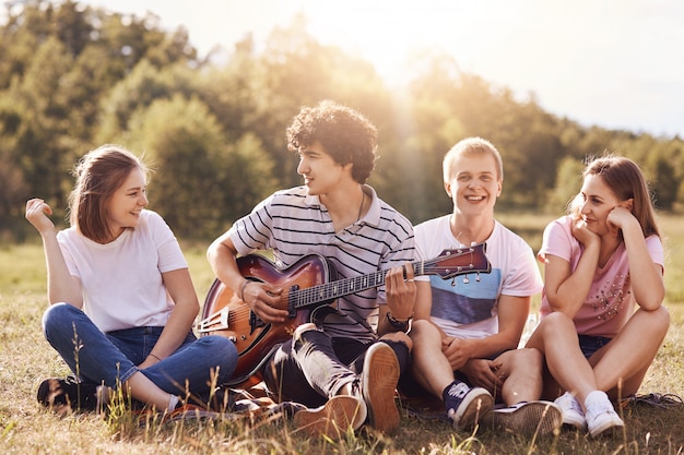 Gelukkige jonge vrienden zingen liedjes voor gitaar, hebben samen vreugde, recreëren buiten, zitten op groen gras. Krullend knappe mannelijke tiener speelt gitaar, enteratins zijn metgezellen, geniet van een warme zomerdag.