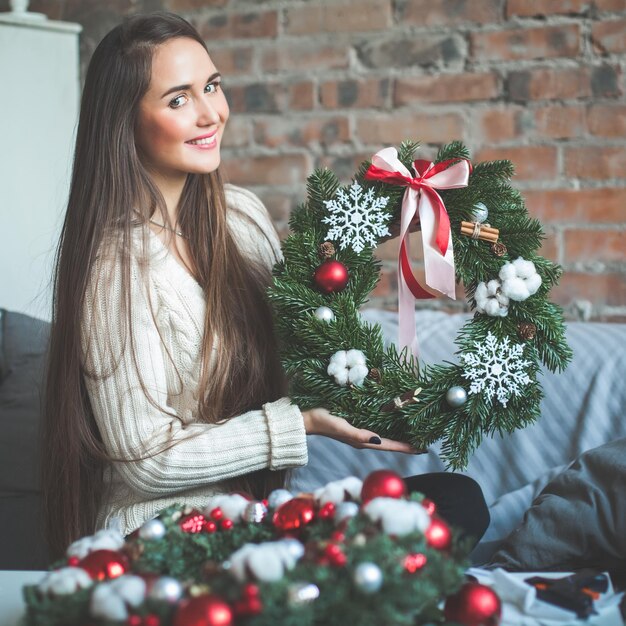 Gelukkige jonge vrouw die de altijdgroene boomkroon van Kerstmis met rode glasballen, kegels, lint en kaneel thuis toont