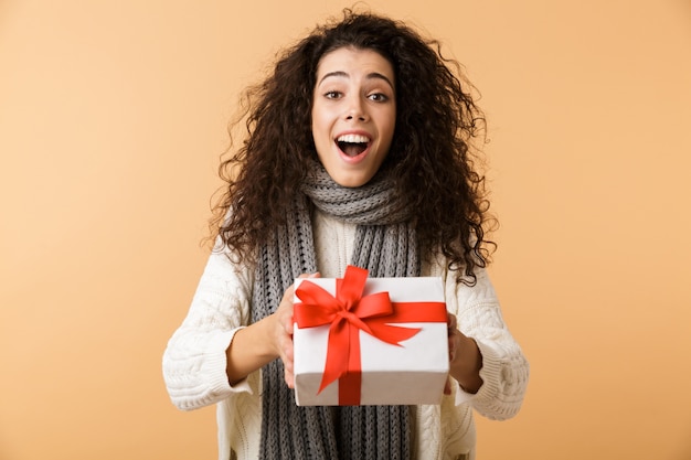 Gelukkige jonge vrouw die de wintersjaal draagt die zich geïsoleerd over beige muur bevindt, die huidige doos houdt