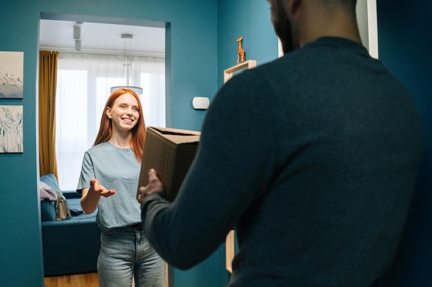 Gelukkige jonge vrouw die een kartonnen doos ontvangt van een onherkenbare bezorger voor de deur van het appartement