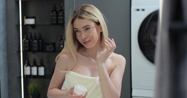 Foto gelukkige jonge vrouw die huidverzorgingscrème op haar gezicht doet kijk in de badkamer spiegel gezichtscrème gezond huidverzorgdheidsbeleid