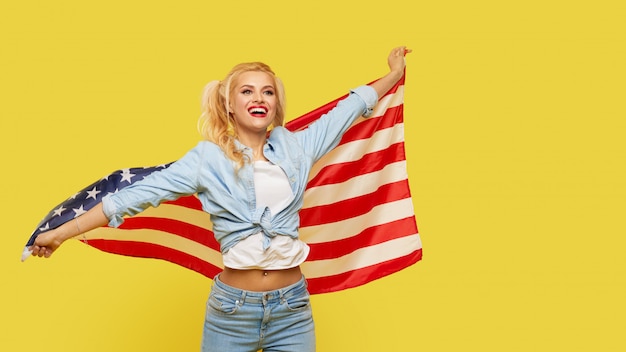Gelukkige jonge vrouw die in denimkleren de vlag van de VS op gele achtergrond houden.
