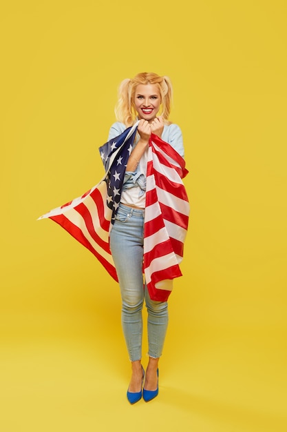 Gelukkige jonge vrouw die in denimkleren de vlag van de VS op gele achtergrond houden.