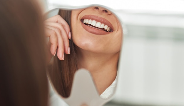 Foto gelukkige jonge vrouw die lacht en haar perfecte gezonde tanden in de spiegel van dichtbij bekijkt, op het kantoor van de tandarts.