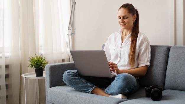 Gelukkige jonge vrouw die laptop thuis met behulp van. Op de bank naast de vrouw staat een camera. Fotograaf retoucheert foto's thuis.