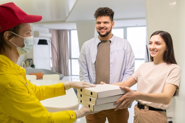 Gelukkige jonge vrouw die met een brede glimlach naar een mannelijke koerier kijkt in handschoenen en een beschermend masker dat haar stapel dozen met bestelde pizza passeert