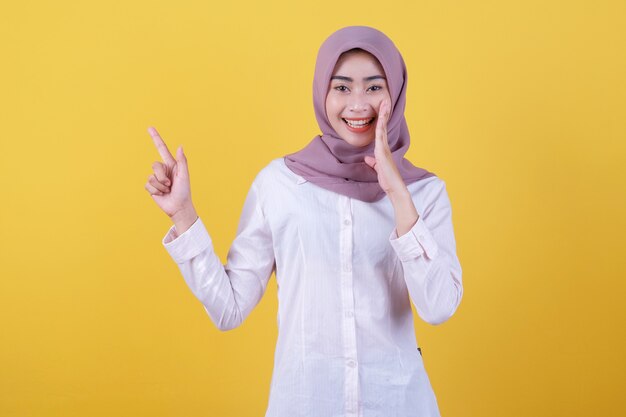 Gelukkige jonge vrouw die met haar vinger naar links wijst en hijab draagt en fluisterde iets