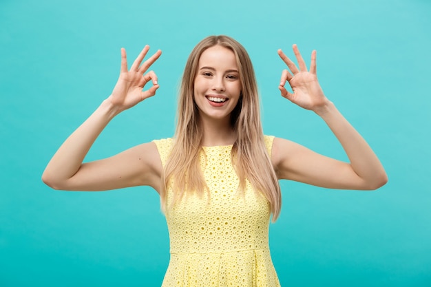 Gelukkige jonge vrouw die ok teken met vingers met grote glimlach toont die op een blauw wordt geïsoleerd