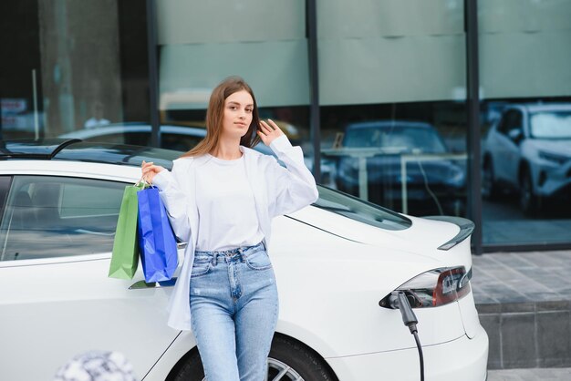Gelukkige jonge vrouw die op stadsparking staat in de buurt van een elektrische auto die de auto-accu oplaadt van een klein stadsstation met boodschappentassen