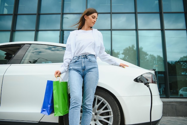 Gelukkige jonge vrouw die op stadsparking staat in de buurt van een elektrische auto die de auto-accu oplaadt van een klein stadsstation met boodschappentassen