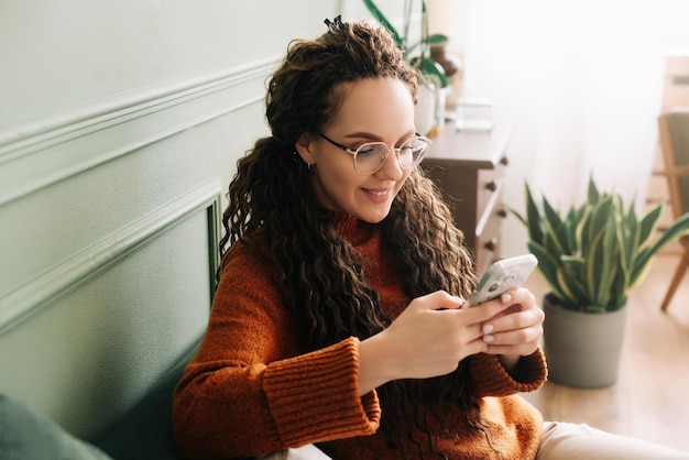 Gelukkige jonge vrouw die smartphone gebruikt voor online winkelen op de bank thuis Ontspannen dame die geniet van technologie en mobiele apps voor e-commerce