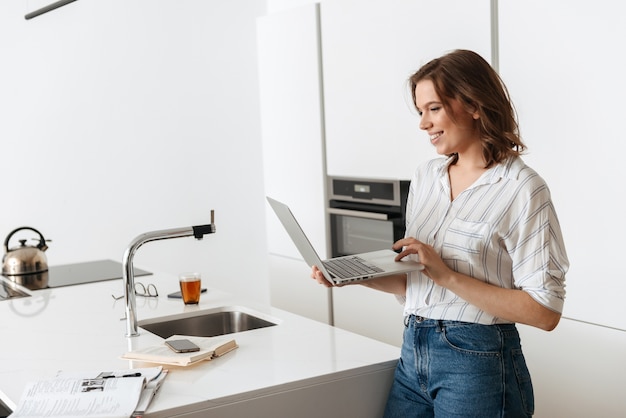 Gelukkige jonge vrouw die zich bij de keuken thuis bevindt, die op laptop computer werkt