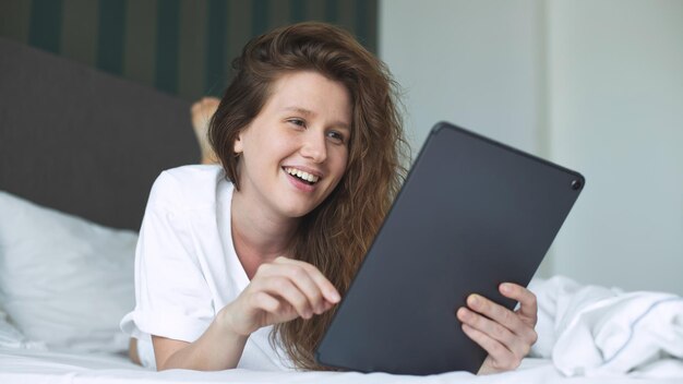 Gelukkige jonge vrouw gebruikt tablet computer thuis in bed in slaapkamer glimlach plezier hebben