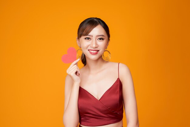 Gelukkige jonge vrouw gekleed in rode jurk met papieren hart geïsoleerd op gele achtergrond
