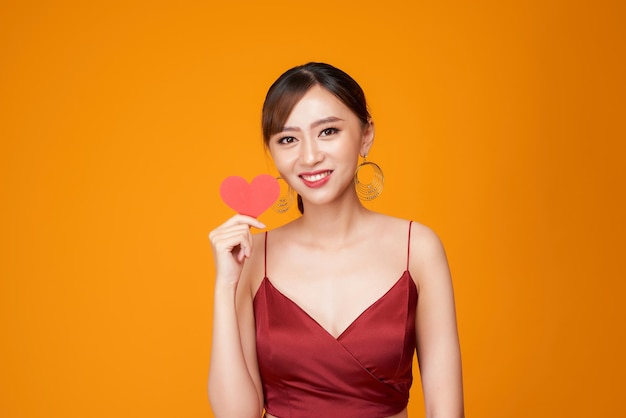 Gelukkige jonge vrouw gekleed in rode jurk met papieren hart geïsoleerd op gele achtergrond