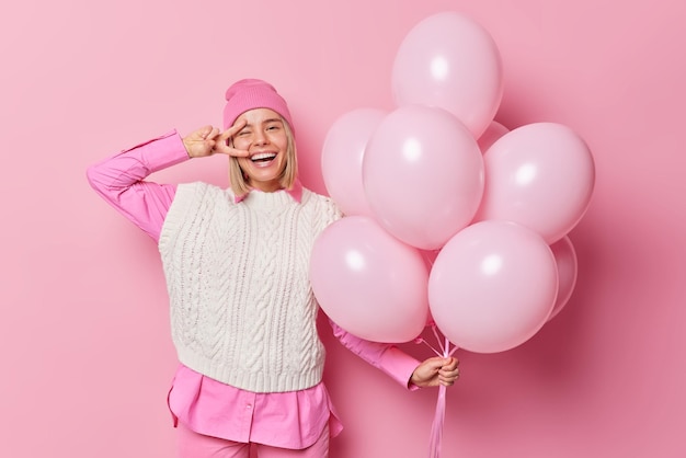 Gelukkige jonge vrouw glimlacht breed maakt vredesgebaar over oog draagt hoed shirt en wit gebreid vest heeft feestelijke stemming komt op kippenfeest poses met bos ballonnen tegen roze achtergrond
